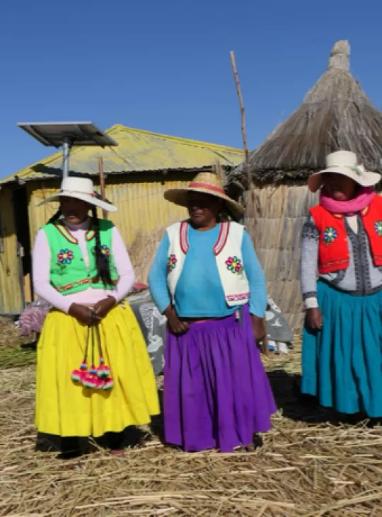 Drie Peruaanse dames staan in kleurrijke kleding voor hun rondavels